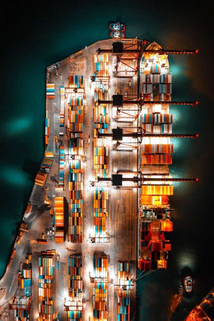 Dramatic aerial view of a busy shipping port with colorful containers and bright lights at night.